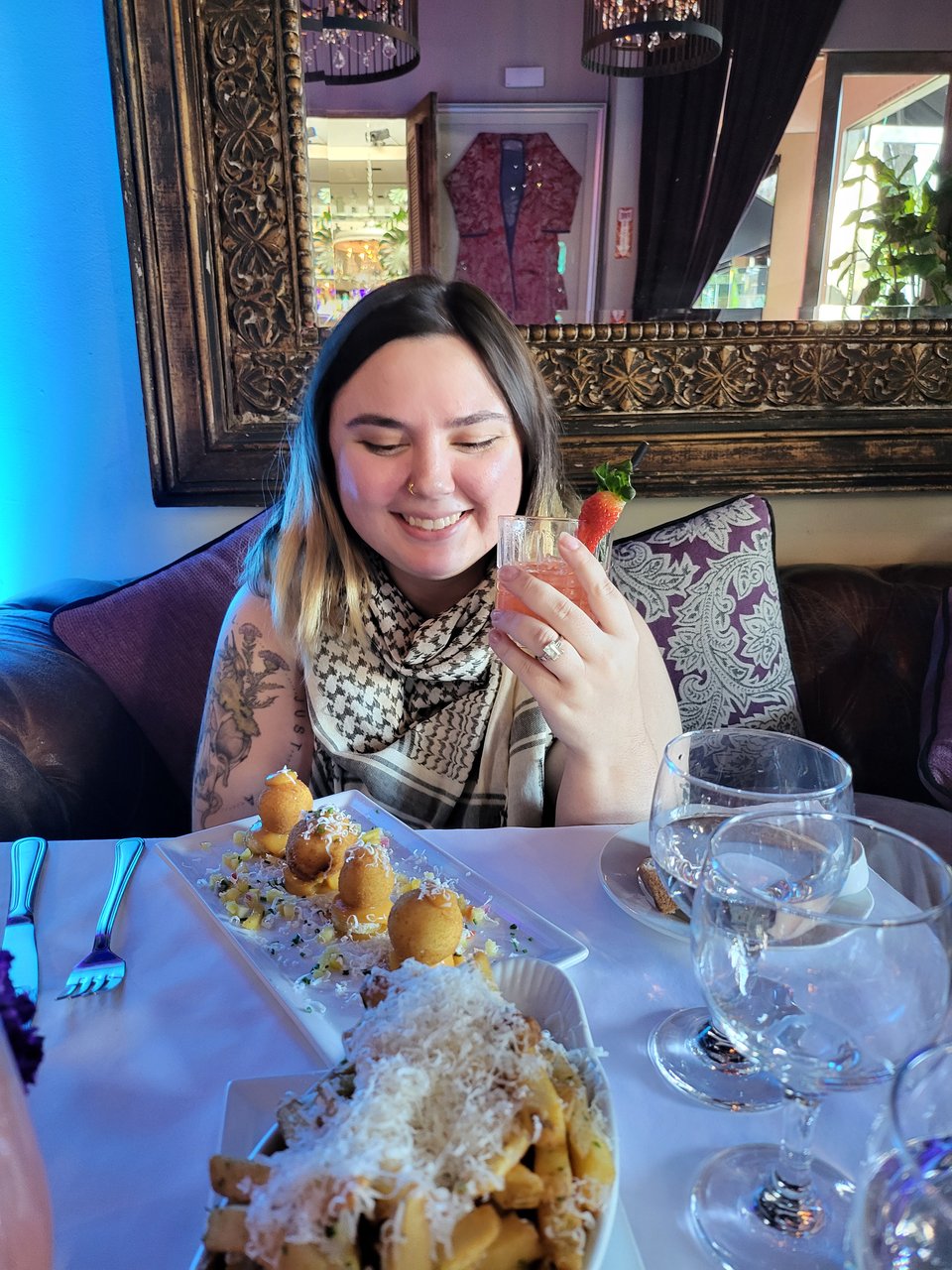 Becca sits smiling at a table at the SUR restaurant in L.A. There are truffle fries and goat cheese balls in front of them. They've got a pink drink in their hand (and are casually showing off their engagement ring) and there's a big mirror behind them.
