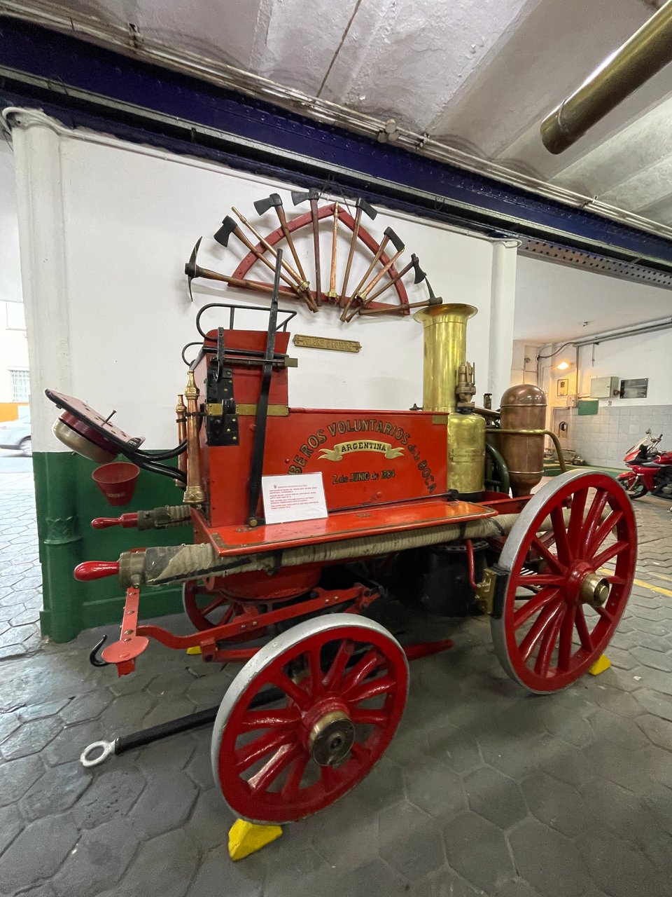 A steam engine fire pump from 1884 on display with eight axes formed in an arch on display on the wall behind the engine.