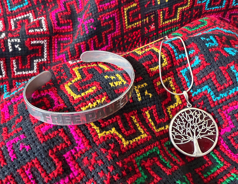 Photograph of a silver cuff bracelet engraved with "FREE PALESTINE" and a silver round pendant necklace featuring a tree design, both placed on a colorful, patterned textile with red, pink, yellow, blue, and black geometric shapes, which is typical of Palestinian embroidery