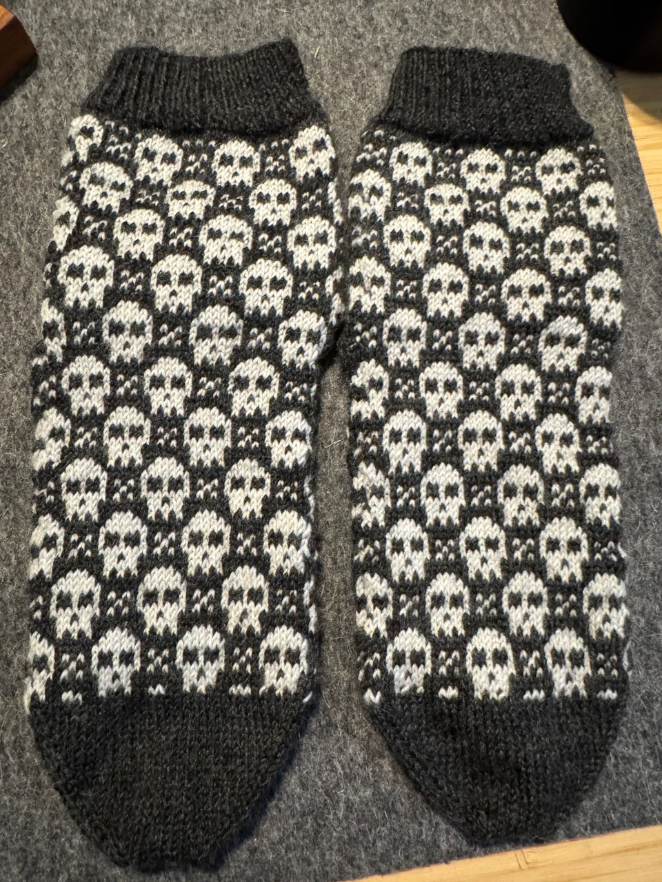 A pair of hand knit socks in gray and white with a repeating pattern of skulls and x shapes.
