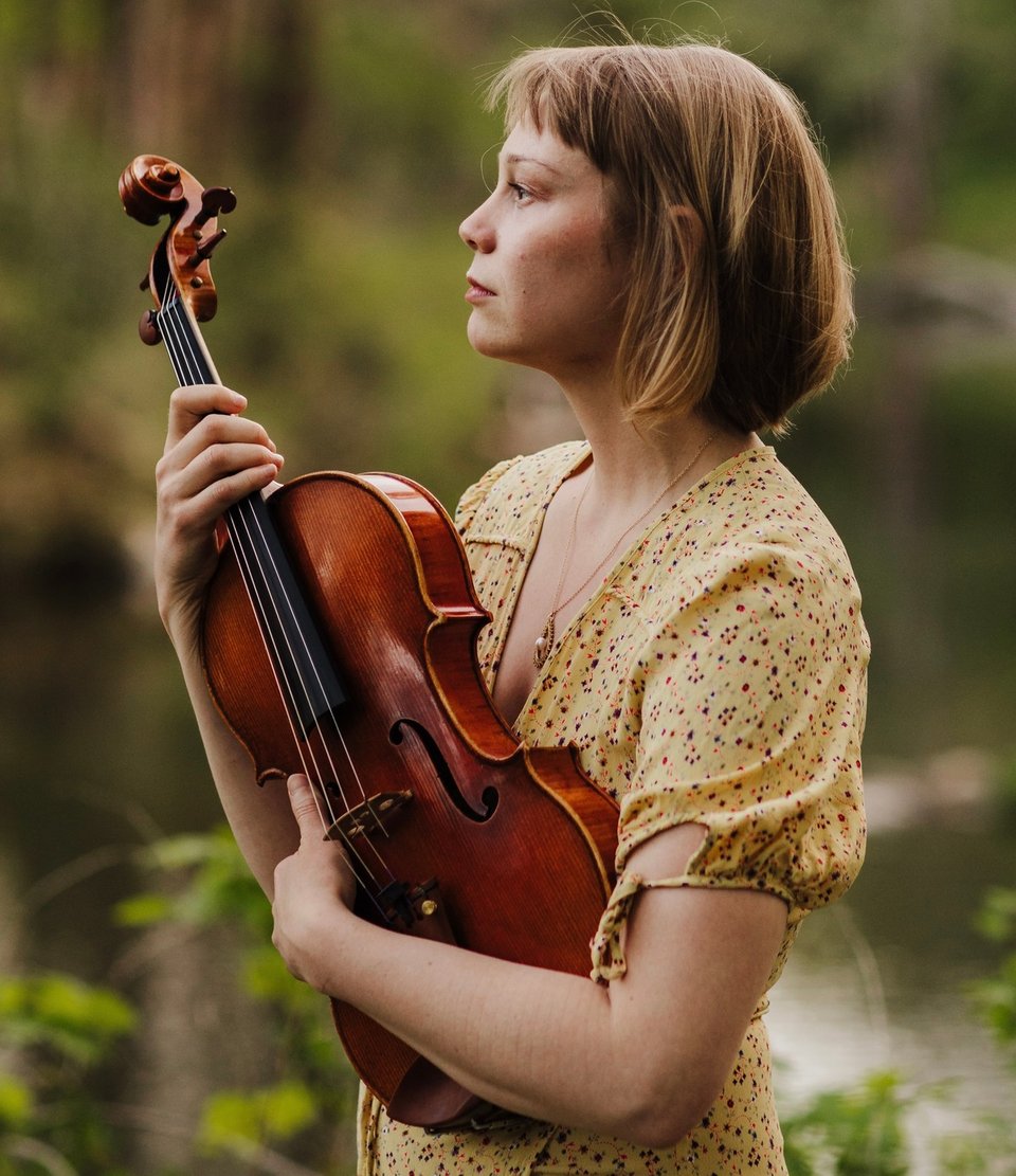 A woman in a yellow print dress with short blond hair cradles a viola. She is shown in profile, facing left.