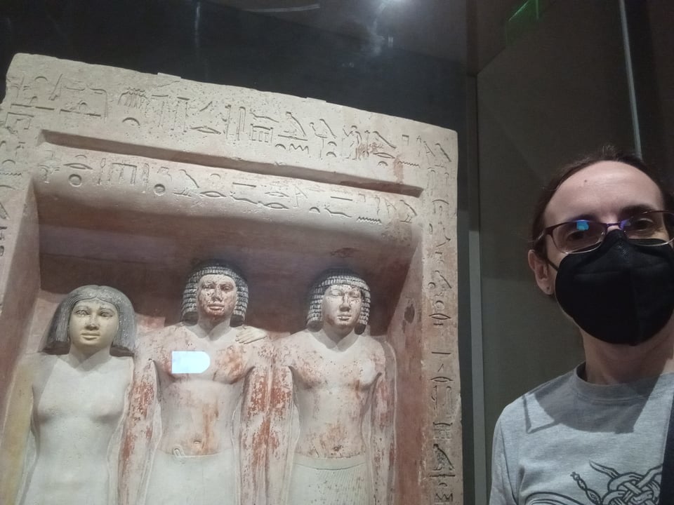 A selfie of me, a white middle-aged lady in a black mask, in front of a threesome of ancient Egyptian statues (2 men and a woman).