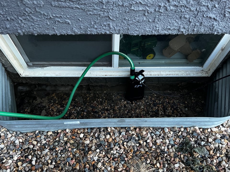 Photo looking down into a basement window well with a pump pumping water out of the well.