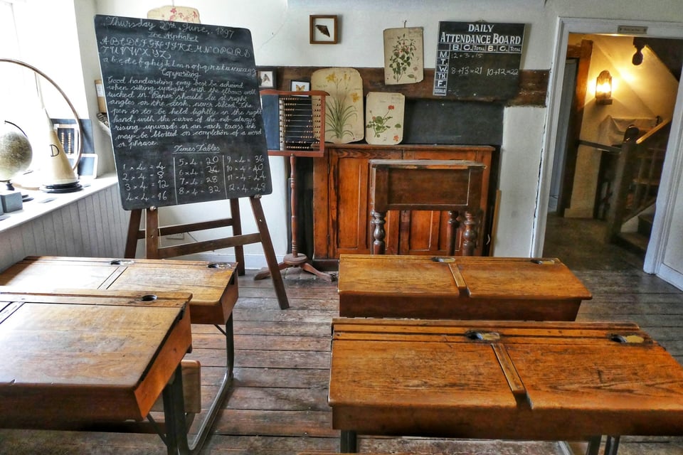 Four wooden desks and a chalkboard, in a setup reminiscent of an old-time classroom.  Photo by Belinda Fewings on Unsplash.