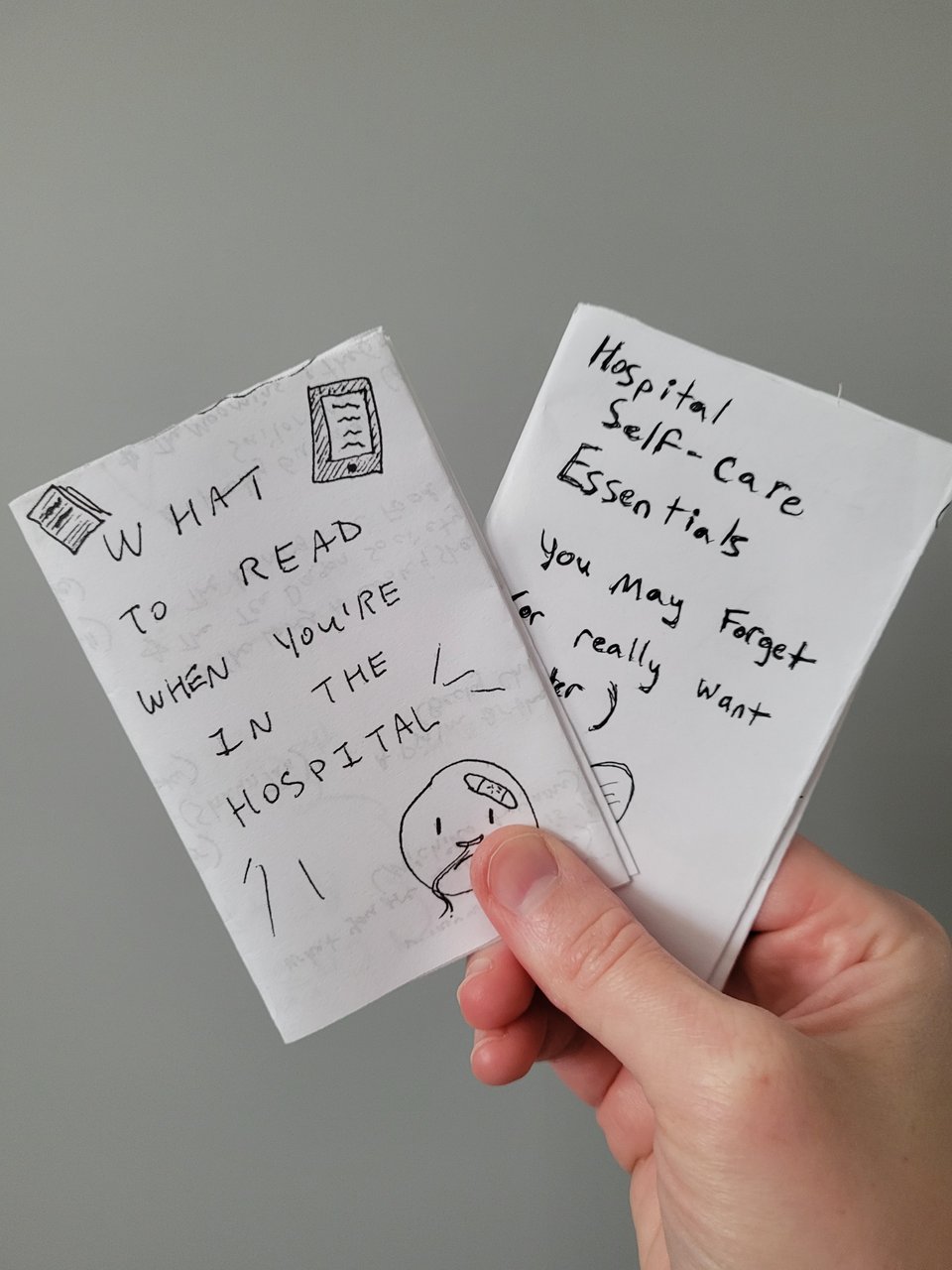 a light-skinned white hand holding two paper minizines. The one on the left is titled "What to read when you're in the hospital", and the one on the right is titled "Hospital self-care essentials you may forget (or really want later)"