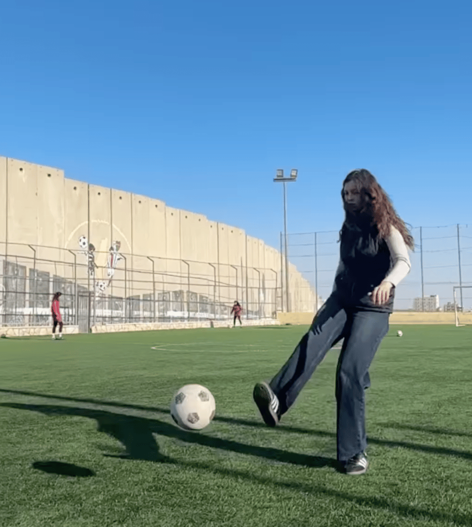 A girl kicking a soccer ball on a field, with Israel's separation wall behind the field