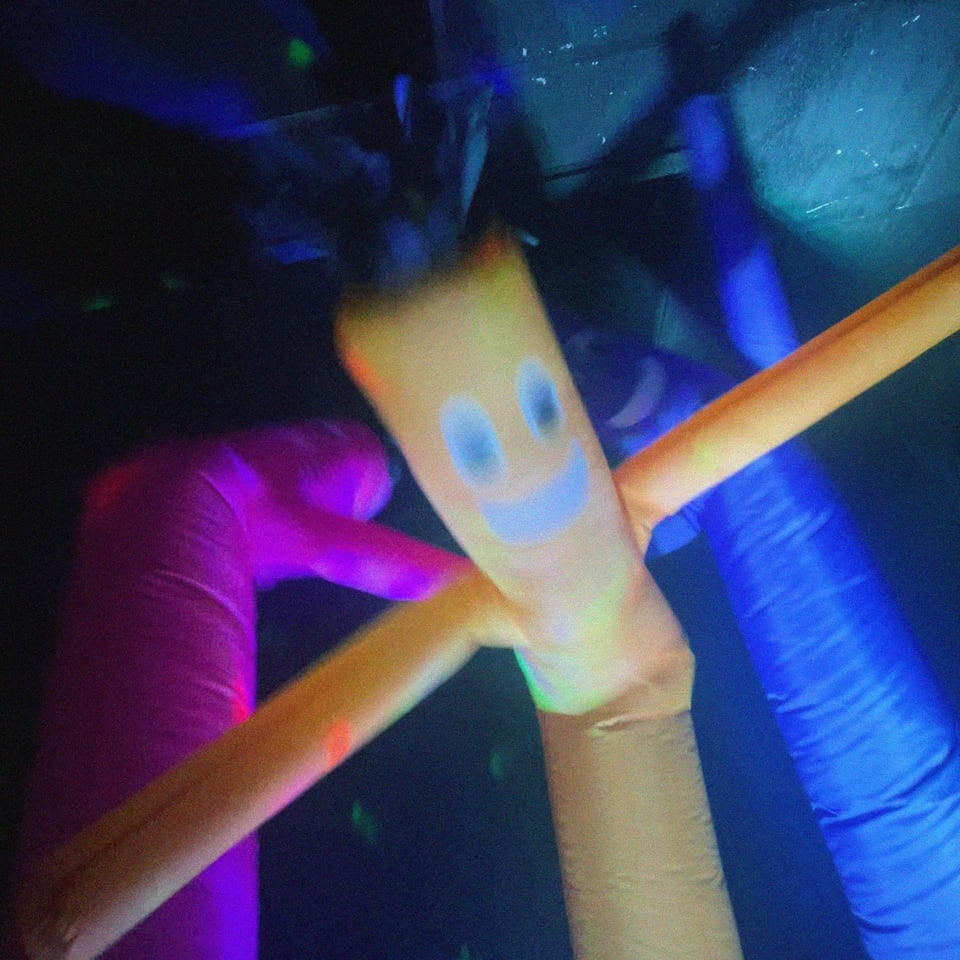 three "wacky wiggly arm guys" aka air dancers (colors: pink, yellow, blue) captured in mid mania, smiling almost threateningly, shot from a low angle like a 90s hip hop video