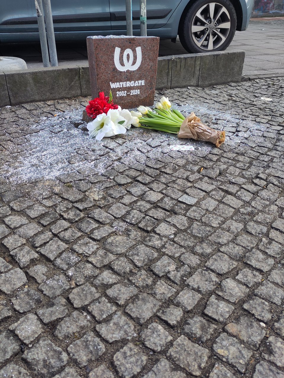 Ein improvisierter Grabstein steht auf Gehwegpflaster. Inschrift: "Watergate, 2002 - 2024". Davor liegen Blumen, es ist ringsum ein wenig Silberglitzer versprüht.
