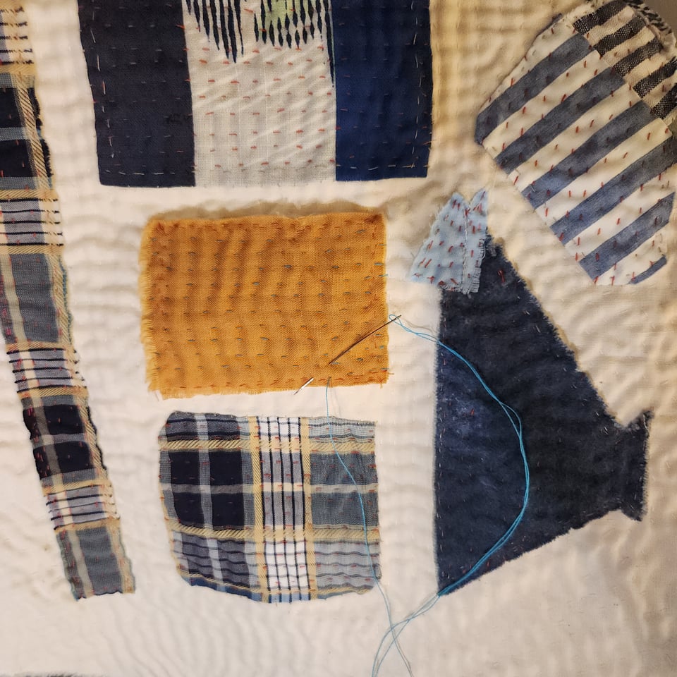 Detail view of Meet Me at the Corner in progress. This view has one piece of dark yellow fabric surrounded by pieces in different shades of blue. A needle and thread are visible.
