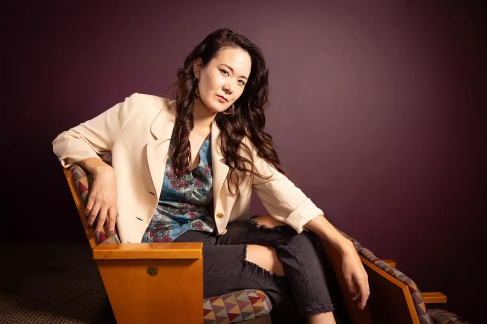 Playwright Keiko Green, wearing a tan jacket and leaning in a theatre seat