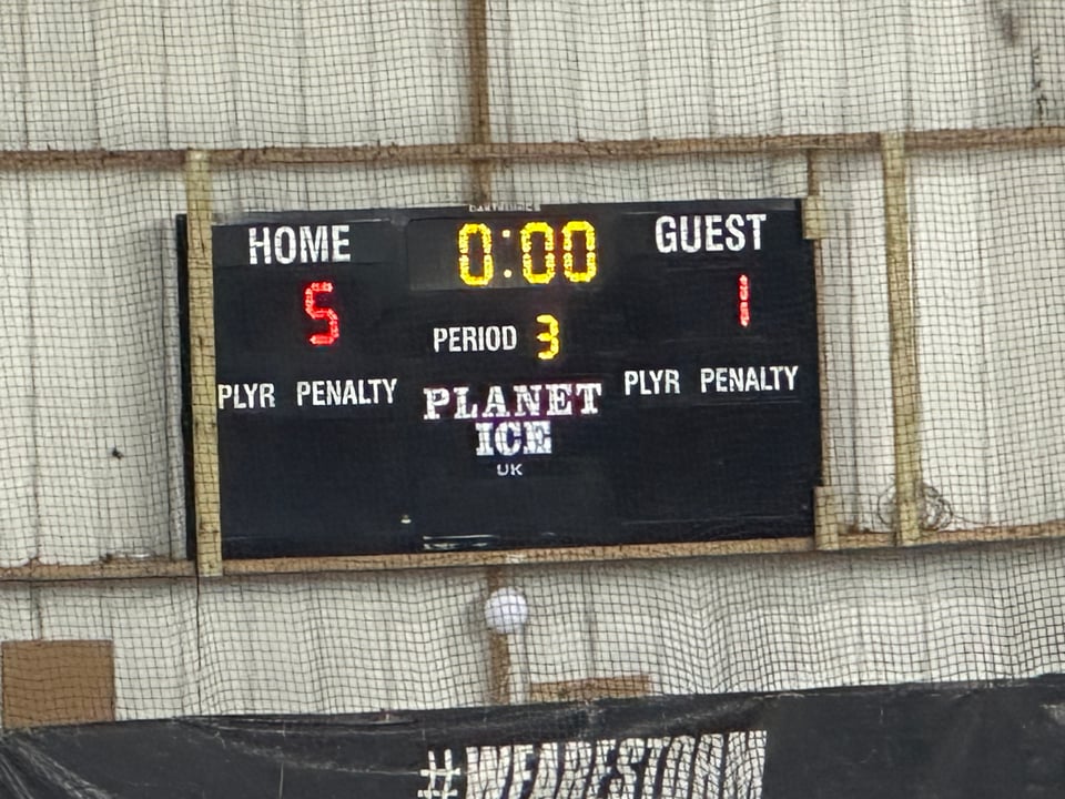 A scoreboard with white, red and yellow text, mounted on a crude looking wall surrounded by netting.