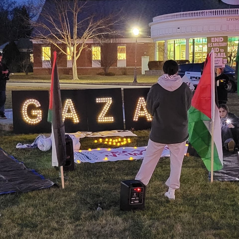 A woman in a keffiyeh stands in front of lit white letters on a black background spelling "GAZA" flanked by Palestinian flags. She is facing the sign, with her back to the camera.