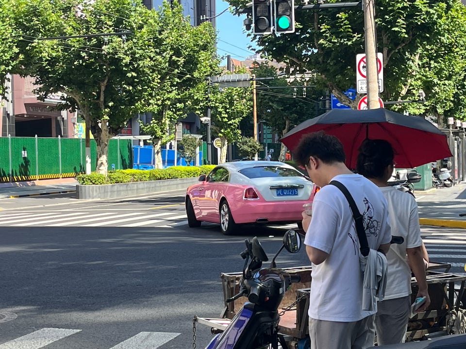 A photo of the car turning at a stoplight, with two people in the foreground standing at the intersection under an umbrella offering shade from the sun.