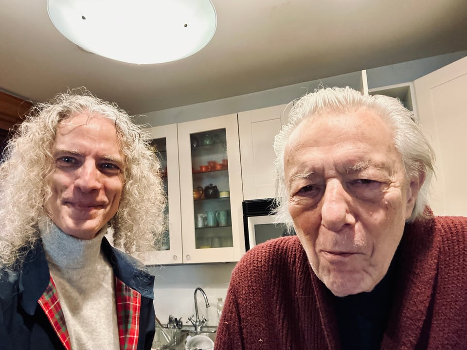 photo of two white men in a kitchen; left is a man in his 50s wearing a light gray turtleneck and navy Harington G9 jacket, with long curly hair coming down to his shoulders; right is the great artist Ed Sorel, mugging for the camera, wearing a red/maroon cardigan over a black shirt. The lighting isn't great, but hey.