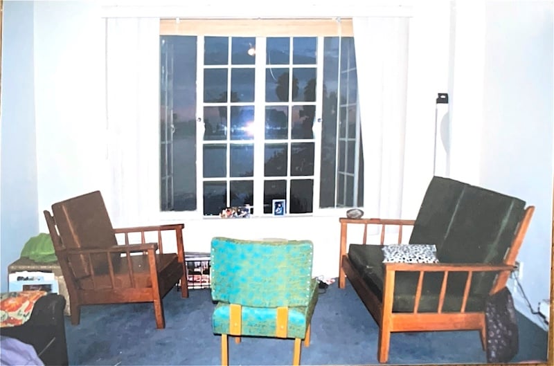 A studio apartment living space with light blue carpet, white walls, wooden furniture and upholstered chair, big windows with palm trees in the distance.