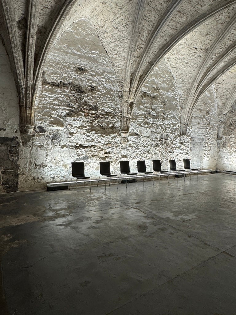An image of St Mary's Abbey in Dublin, Ireland. The Chapter House of the Cistercian monastery is a rib-vaulted chamber, moodily lit from the floor. Seven black chairs (in a contemporary style) are arranged against the wall.