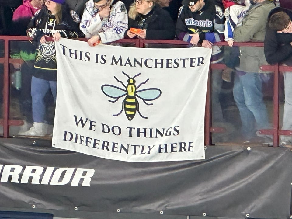 A flag hung over a balcony. Picture of a bee. Text: This is Manchester we do things differently here.