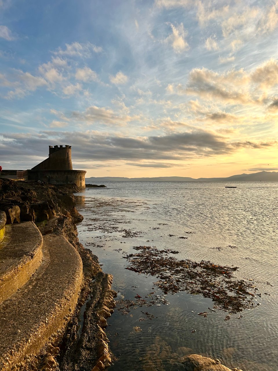 A sea wall in Scotland. Seaweed is floating on the water and the sun is setting over distant island mountains. Image by Rowan Ambrose.