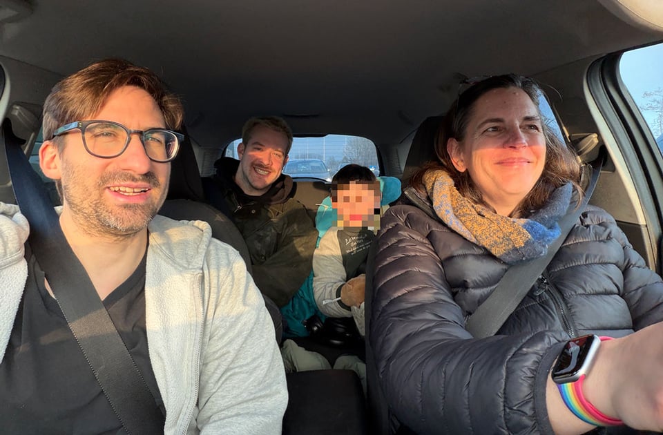 Four people are in a car - two in the front seats, two in the back. All smiling, but the child's face is blurred.