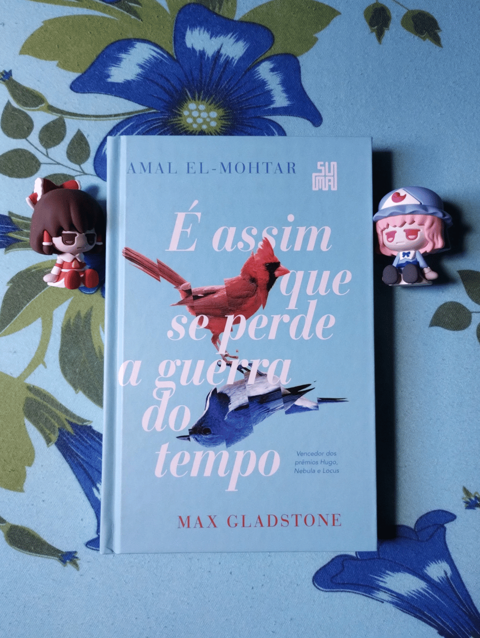 O Livro “É Assim que se Perde a Guerra do Tempo” em um cenário azul com flores, ao lado de duas bonequinhas personagens da série Touhou Project