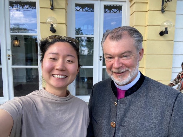 young East Asian woman takes selfie in front of light yellow and white palace front entrance wall with white male bishop