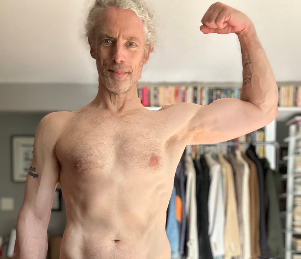 photo of shirtless white man flexing his left bicep and smiling at the camera; he looks okay, and his forearm is kinda swole
