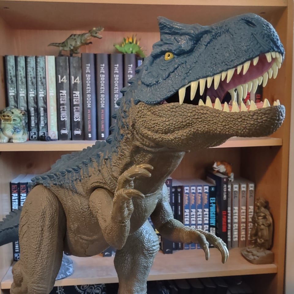A (toy) blue and olive-green Allosaurus looms in front of a bookshelf filled with books by that Peter Clines guy.