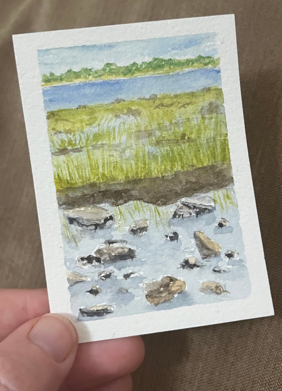 Maggie's hand holds a small painting of rocks in a tide pool surrounded by green beach grass, with the ocean and Plum Island in the background.