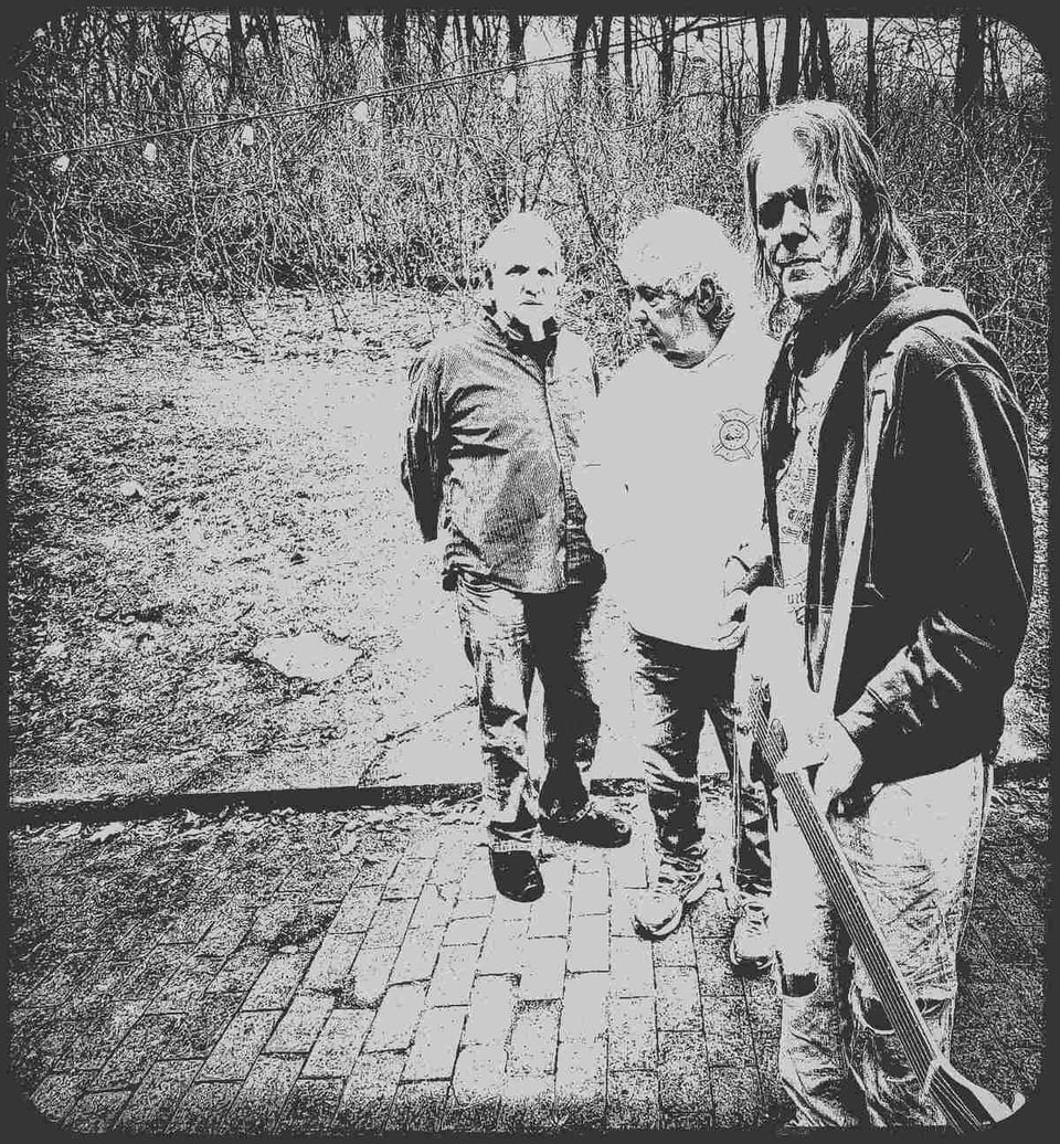Three men stand next to each on a patio made of brick pavers. The one nearest the camera holds an electric guitar. The photo is a black-and-white, high-contrast image that has a washed-out appearance.