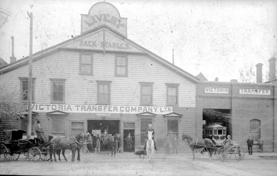 Black and White photo. A large wooden building with a sign that reads "Livery - Hack Stables - Victoria Transfer Company Ltd." There are horses, carriages, and people outfront posing for the photo