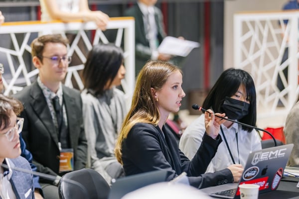 A student speaks into a microphone while seated with other participants at a conference table.