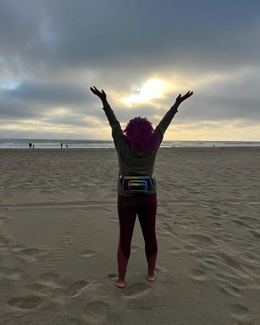 Photo of Patricia from behind at the beach. She is facing the ocean and sunset. Her arms are raised high.
