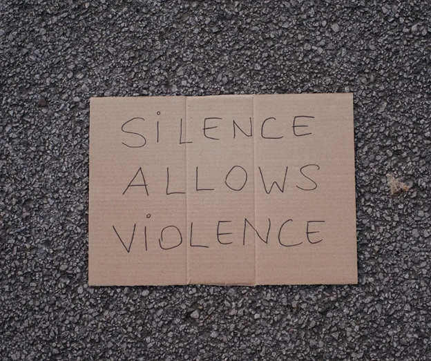 A brown cardboard sign with "SILENCE ALLOWS VIOLENCE" handwritten in all caps with black marker. The sign is displayed on a background of black asphalt.