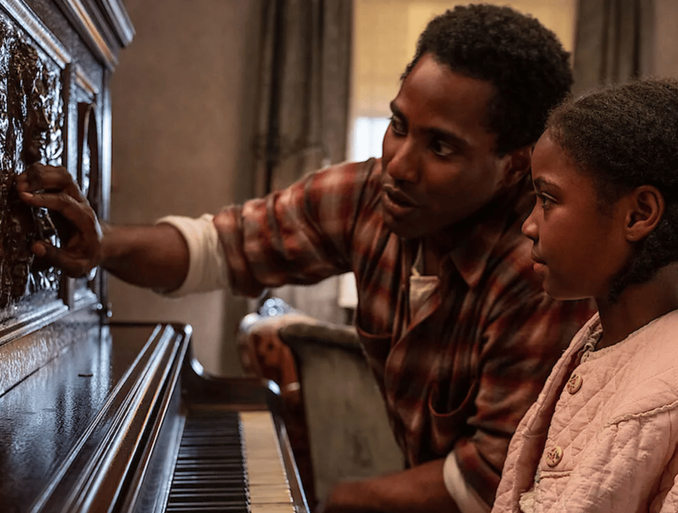 Actors John David Washington and Skylar Aleece Smith sit side by side at an old, brown piano.