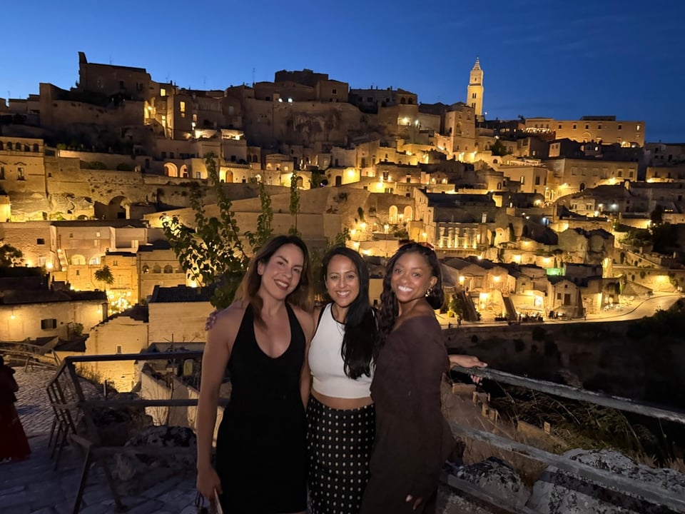 Nadia stands on the right with two friends at a viewpoint overlooking Matera, with the ancient city glowing with golden lights against the deep blue night sky.
