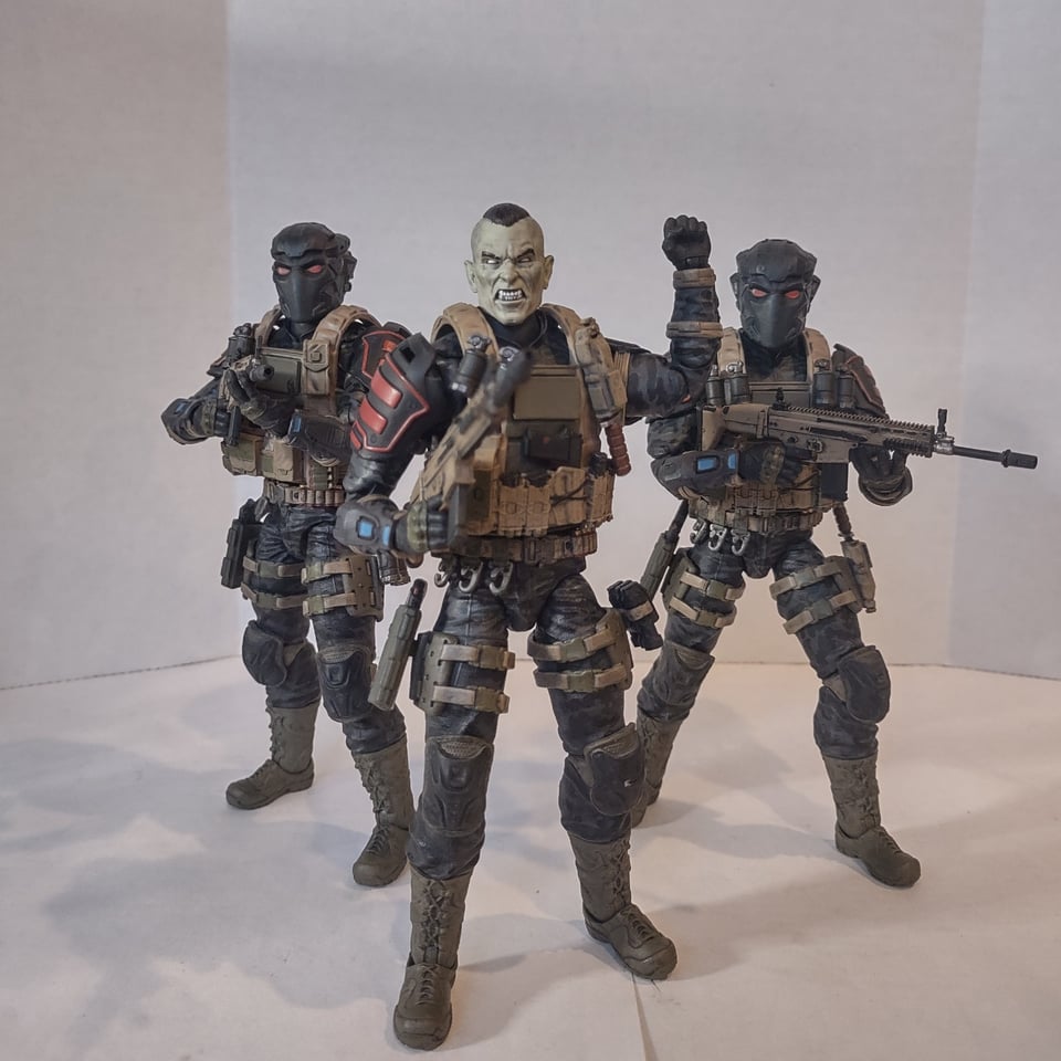 A trio of military action figures in dark fatigues, each wearing body armor loaded with gear and carrying a rifle. The two in the back wear full-head, futuristic helmets with bright red eyes, The one in the foreground, signaling the others with his fist raised, has no helmet, revealing his pale skin and eyes, pointed ears, and fangs.