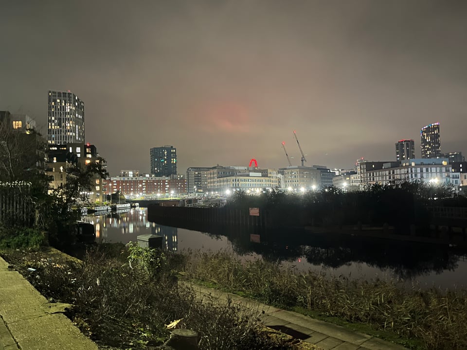 A dark canal with a path along the edge, a few lights, and a lit-up city skyline in the background.