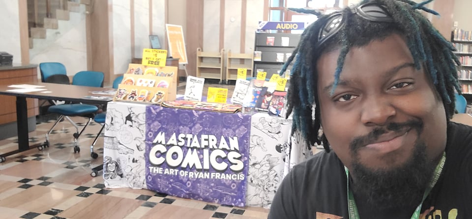Photograph of Ryan"Mastafran"Francis at his table at Fandom, Art, and Music Expo 2026, showing his selection of self-published comics, stickers, and tablemats.