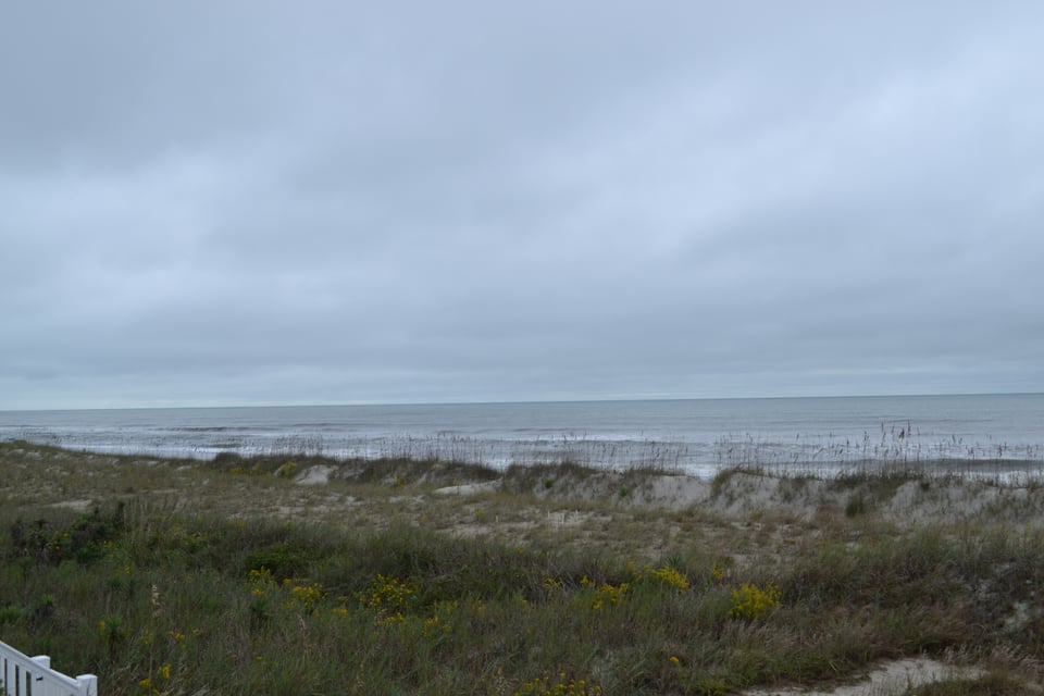 A view of the ocean from low dunes that are made of white sand covered with grass and oats. The sky is a threatening gray.