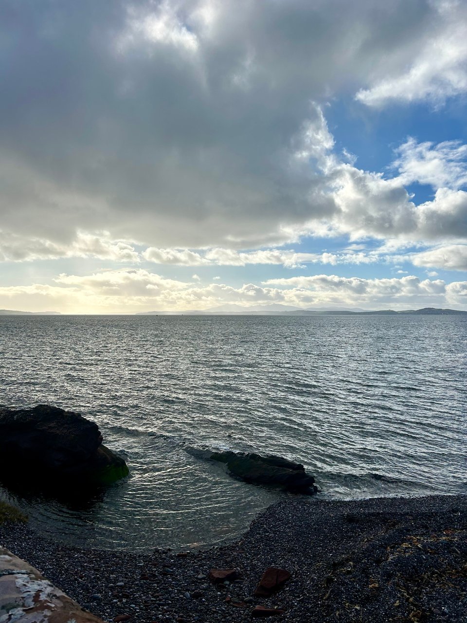 Hazy mountains are in the far distance across the Firth of Clyde in Scotland. The water is glittering grey, there are patches of blue sky and lots of dark clouds. Image by Rowan Ambrose.