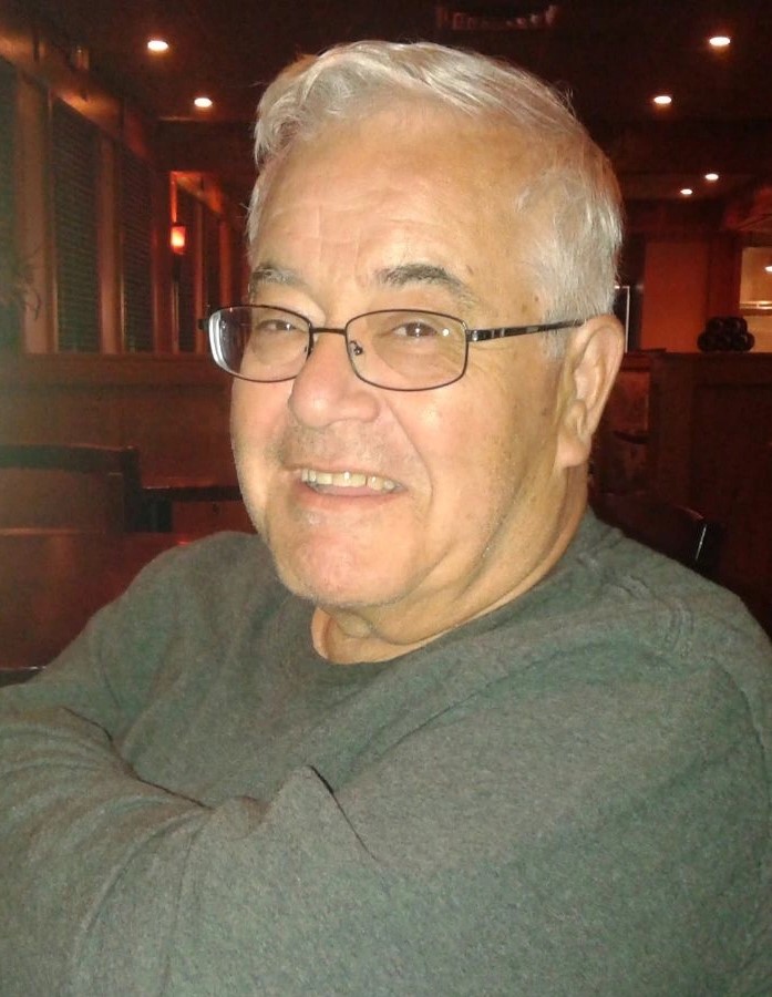 My father, a white man with grey hair and blackish eyebrows who is wearing glasses and smiling at the camera while crossing his arms over his chest.