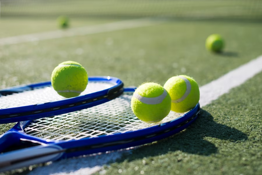 Two blue tennis rackets on the ground, with three tennis balls on them. In the distance can be seen two more tennis balls