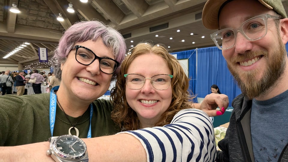 Three smiling people with glasses take a selfie in a cement convention hall.