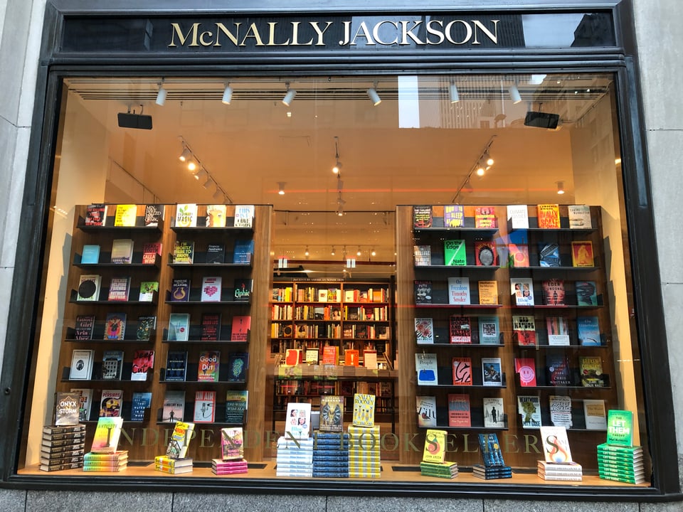 Window of McNally Jackson bookstore with tall bookcases and a view into the store in between them. HUMANS is in the right-hand window bookcase, second row from the top, third from the left.