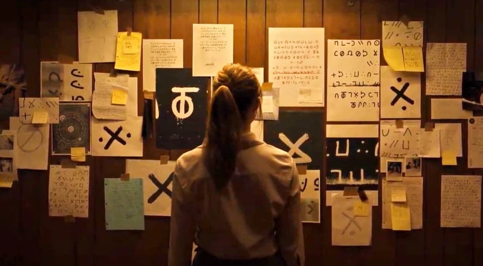 A still from the movie Longlegs (2024). A female with a ponytail stands in front of a wall with a bunch of ciphers on it.