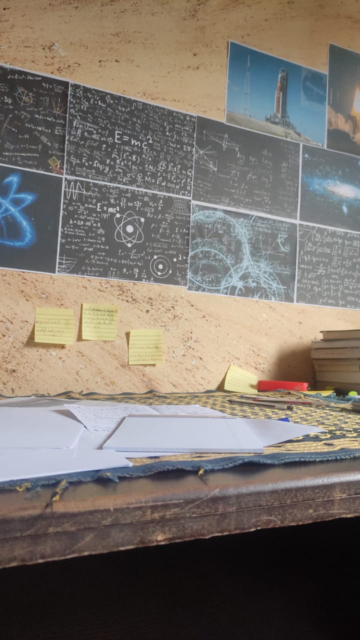 a desk with some papers on it, up against a wall with print outs featuring physics equations and also images from space launches