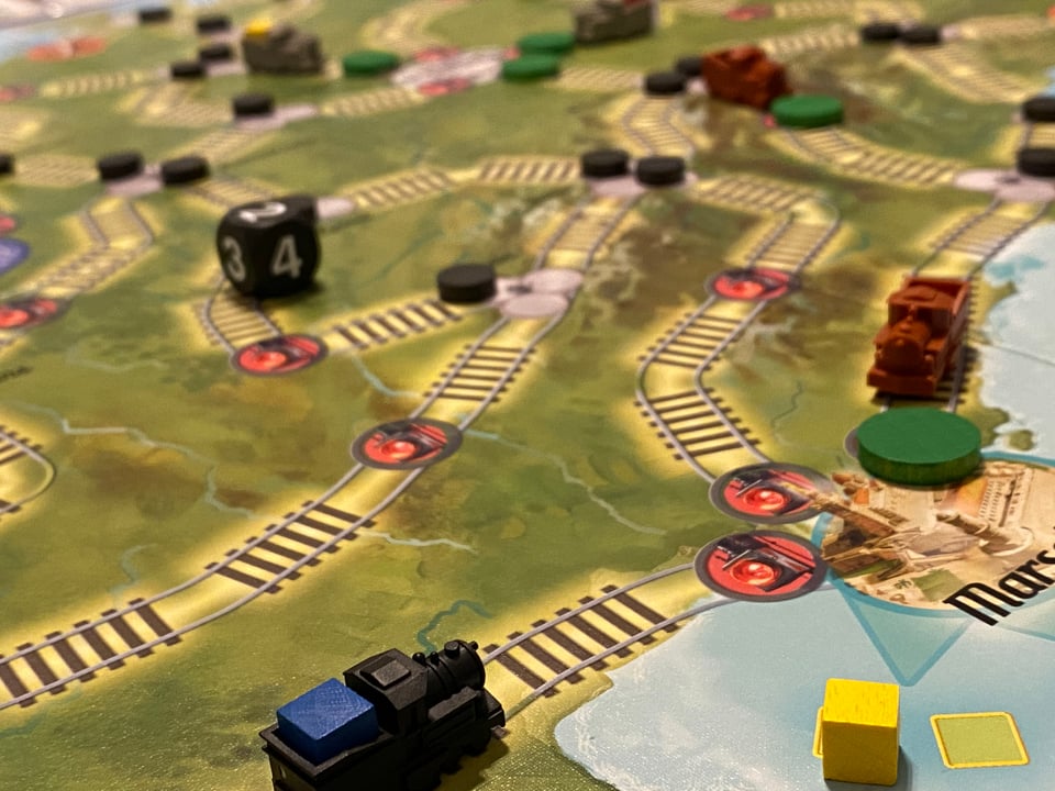 A game board showing train tracks. Plastic train engines carrying wooden cubes and a die are on the board.