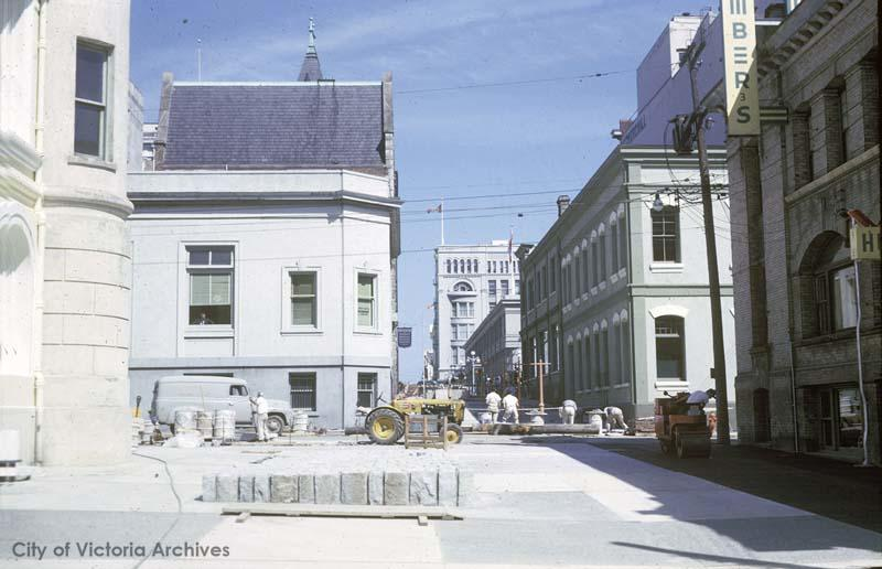 Colour photo. Looking up a streetscape with buildings on all sides. The old court house is to the left. There are workers and a road roller and tractor in the middle of the photo.