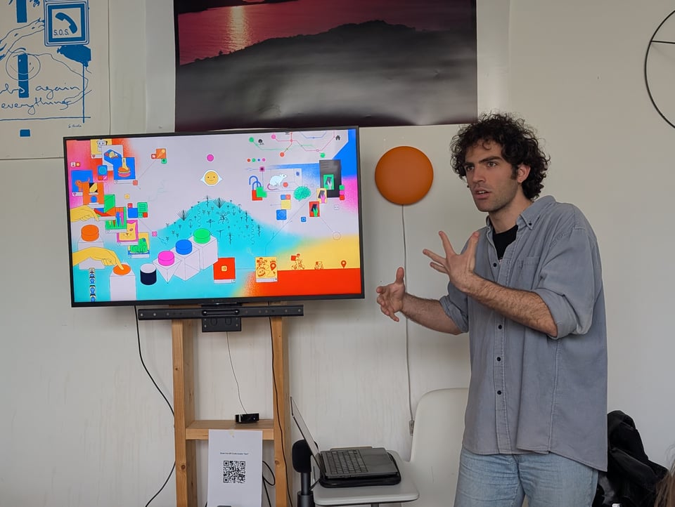A speaker gestures as they speak to an audience. The screen next to them has a colourful pattern on it.