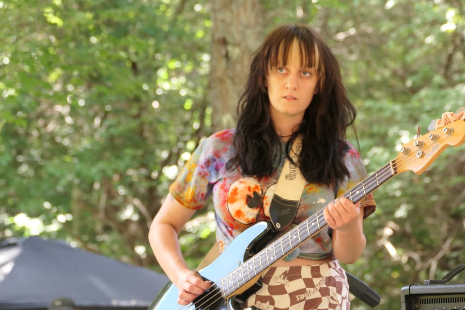 A woman with long brown hair and a colorful print top plays an electric bass guitar.